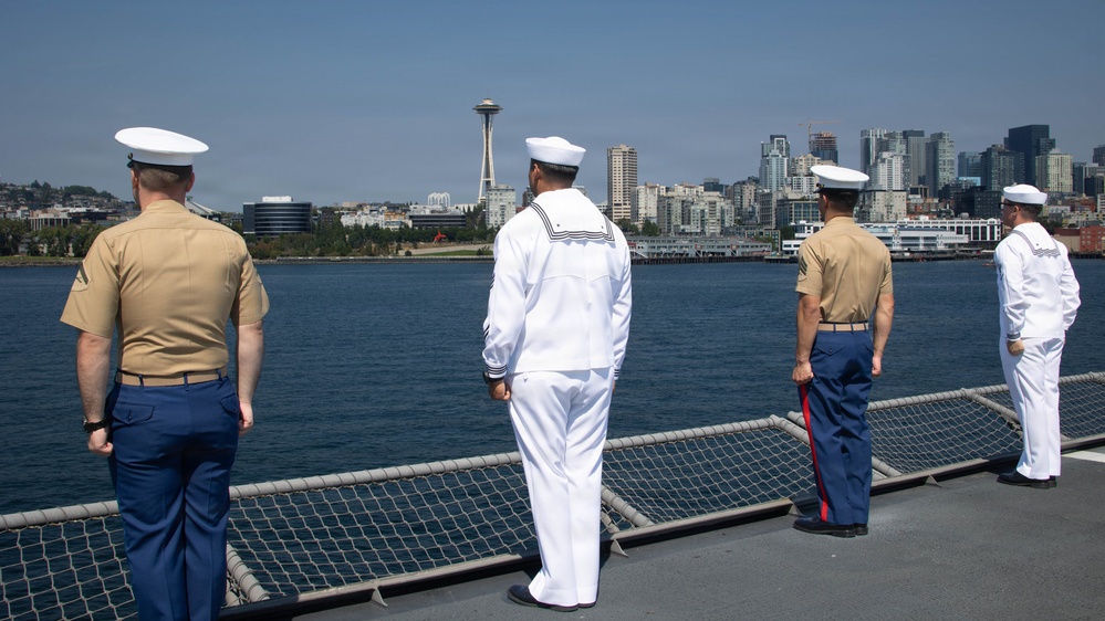 Four military personnel stand at the railing of a naval vessel looking toward a city skyline across the water. The Space Needle is clearly visible in the background, identifying the location as Seattle, Washington. The personnel are wearing a mix of U.S. Marine Corps and U.S. Navy dress uniforms, suggesting a formal occasion such as a fleet week or port arrival.