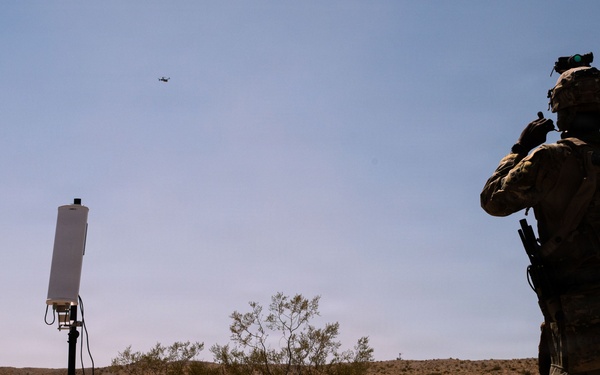 A military or law enforcement operator in full tactical gear stands in an arid desert environment, observing an airborne drone in the sky. A tripod-mounted antenna or sensor panel is positioned nearby, suggesting a counter-drone or communications operation. The setting appears to be a training exercise or field demonstration in a desert scrubland terrain.