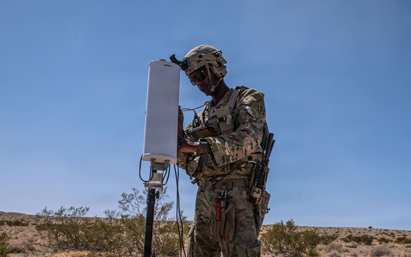 A soldier in full combat gear and camouflage uniform is operating or configuring a communications or sensor antenna mounted on a tripod in an arid desert environment. The equipment appears to be a directional antenna or radar unit connected by cables, likely to a power source visible in the background. A second person and what appears to be a portable generator are visible in the mid-ground.