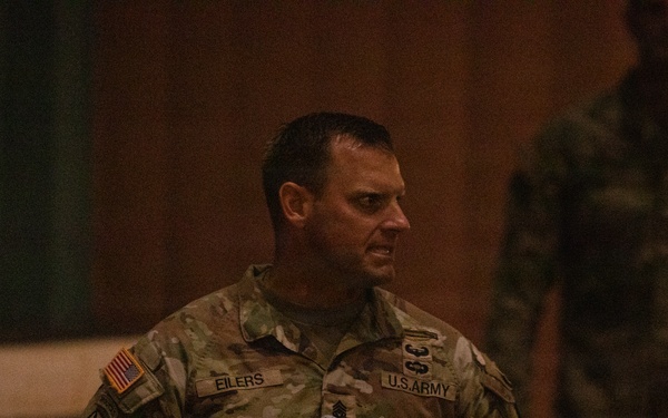A U.S. Army soldier identified by name tape as 'EILERS' is speaking into a handheld microphone, apparently addressing an audience in what appears to be an auditorium or meeting hall. He is wearing the Army Combat Uniform (ACU) in MultiCam/OCP pattern with visible unit patches and rank insignia. A second uniformed individual is partially visible in the background.