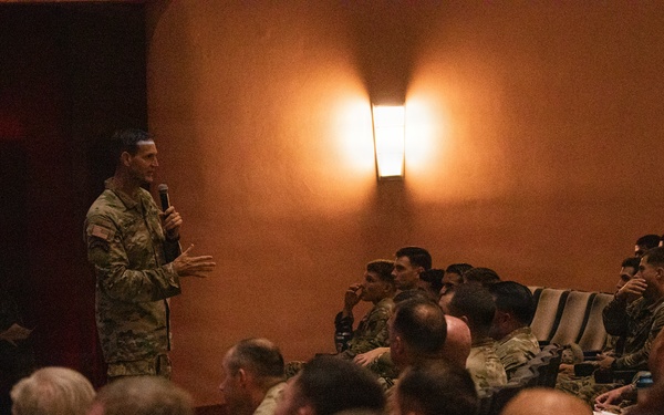 A military briefing or town hall is taking place in a dimly lit room, with one uniformed soldier standing at the front holding a microphone and addressing a seated audience of fellow soldiers. The audience, wearing camouflage uniforms consistent with U.S. Army OCP (Operational Camouflage Pattern), is attentively listening. The setting appears to be an indoor auditorium or meeting room with warm ambient lighting.