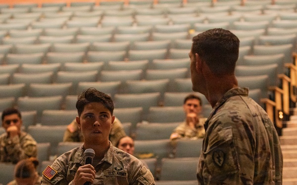 A U.S. Army soldier is holding a microphone and speaking to a senior officer during what appears to be a town hall or Q&amp;A session in a military auditorium. The audience consists of multiple uniformed soldiers seated in tiered theater-style seating. The setting suggests a formal military briefing or command engagement event.