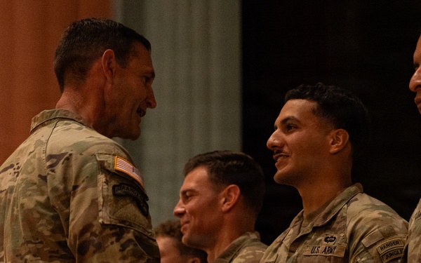 A group of U.S. Army soldiers in OCP (Operational Camouflage Pattern) uniforms are gathered in what appears to be an indoor ceremony or formal event. A senior soldier on the left is shaking hands or presenting something to a soldier in the center, who is smiling. The atmosphere appears positive and ceremonial in nature.