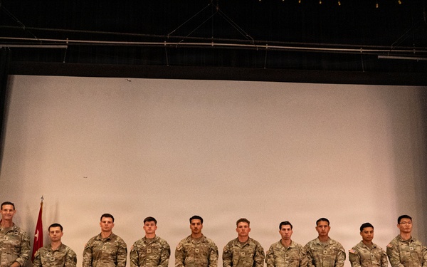 A group of ten military personnel in U.S. Army camouflage uniforms (OCP pattern) are standing in a formal line on a stage, posing for an official group photograph. Two flags are visible to the left, including what appears to be a U.S. flag and a military unit guidon. The setting appears to be an auditorium or theater with a large projection screen backdrop.