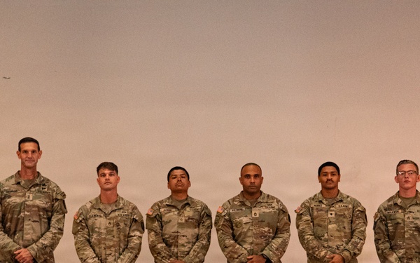 Six U.S. Army soldiers in OCP (Operational Camouflage Pattern) uniforms are standing in a formal line, likely for an official ceremony or recognition event. They are positioned in front of a stage or backdrop with a military flag visible on the left side. All soldiers are standing at ease with hands clasped in front of them.