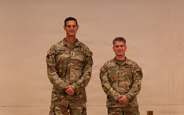 Two U.S. Army soldiers in Operational Camouflage Pattern (OCP) uniforms are standing side by side, posing for a formal photograph. Both wear name tapes and U.S. Army tapes on their uniforms, along with American flag patches on their right shoulders. They are standing on what appears to be a stage or platform in front of a light-colored backdrop.