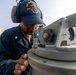 A U.S. Navy sailor wearing a USS Michael Murphy (DDG) ship's cap and hearing protection is looking through a gyrocompass or pelorus navigation instrument aboard a naval vessel. The sailor is wearing Navy working uniform (NWU) with what appears to be a Petty Officer insignia on the collar. The image is taken on the open deck of a destroyer-class warship.