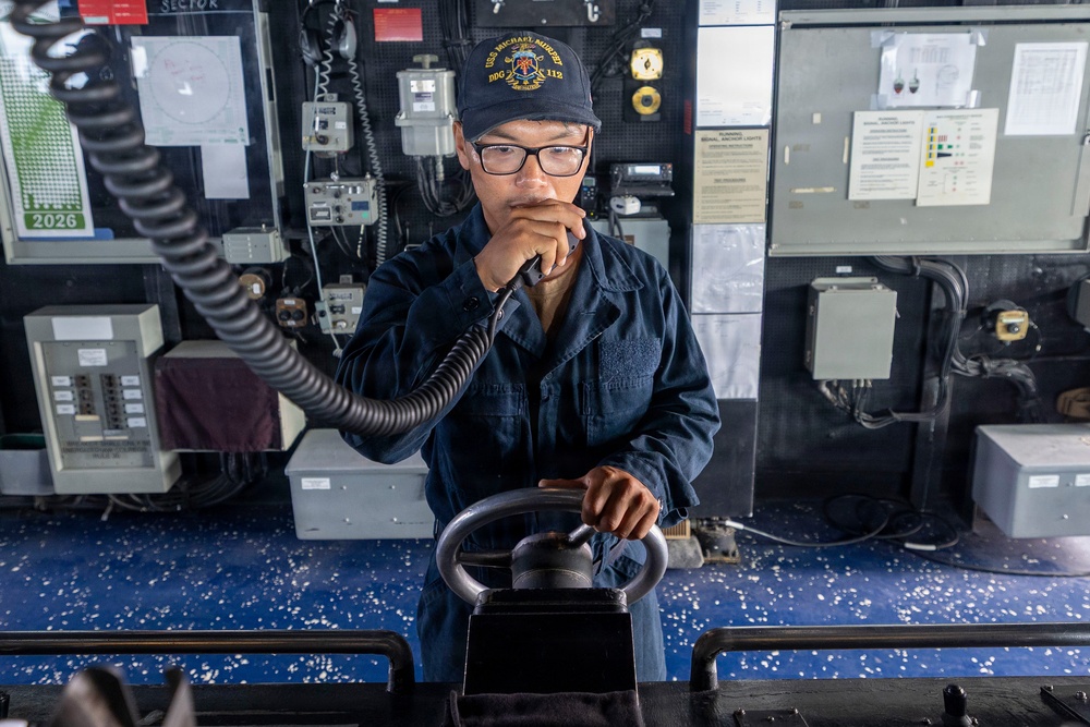 A U.S. Navy sailor stands at the helm of a naval vessel, holding a communication handset while gripping the ship's steering control. The sailor is wearing a Navy working uniform (NWU) and a cap bearing the insignia of USS Michael Murphy (DDG-112). The bridge environment is filled with navigation and communication equipment typical of a guided-missile destroyer.