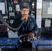 A U.S. Navy sailor stands at the helm of a naval vessel, holding a communication handset while gripping the ship's steering control. The sailor is wearing a Navy working uniform (NWU) and a cap bearing the insignia of USS Michael Murphy (DDG-112). The bridge environment is filled with navigation and communication equipment typical of a guided-missile destroyer.