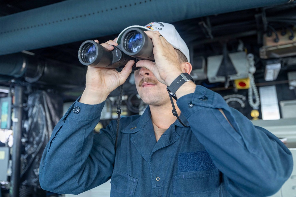A male sailor or naval personnel is using large binoculars to observe something, likely from the bridge or deck of a naval vessel. He is wearing a navy blue coverall uniform and a white cap with what appears to be the number '112' visible. The industrial background with pipes, cables, and equipment confirms a shipboard environment.