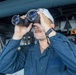 A male sailor or naval personnel is using large binoculars to observe something, likely from the bridge or deck of a naval vessel. He is wearing a navy blue coverall uniform and a white cap with what appears to be the number '112' visible. The industrial background with pipes, cables, and equipment confirms a shipboard environment.
