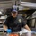 A U.S. Navy culinary specialist is working in a ship's galley kitchen aboard the USS Michael Murphy (DDG-112), ladling red tomato sauce over cooked chicken pieces in a large stainless steel pan. The sailor is wearing a black chef's uniform with the ship's insignia patch, a USS Michael Murphy DDG-112 ball cap, blue nitrile gloves, and hearing protection mounted on the cap. The setting is a professional naval galley with stainless steel equipment visible in the background.