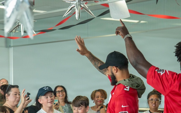Two individuals wearing Atlanta Falcons NFL jerseys (one labeled 'WALKER 11') are interacting with a crowd of fans, including children, in what appears to be an indoor event space. The atmosphere is celebratory, with red and black streamers and decorations hanging from the ceiling. Fans are taking photos and engaging enthusiastically with the jersey-wearing individuals.