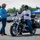 A motorcycle safety or skills training course is taking place in an open parking lot area. An instructor wearing a blue shirt with a lanyard is gesturing and speaking to a student seated on a black Harley-Davidson motorcycle. Orange traffic cones mark the course boundaries, and a blue canopy tent bearing what appears to be an Air Force logo is visible in the background.