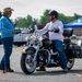 A motorcycle safety or skills training course is taking place in an open parking lot area. An instructor wearing a blue shirt with a lanyard is gesturing and speaking to a student seated on a black Harley-Davidson motorcycle. Orange traffic cones mark the course boundaries, and a blue canopy tent bearing what appears to be an Air Force logo is visible in the background.
