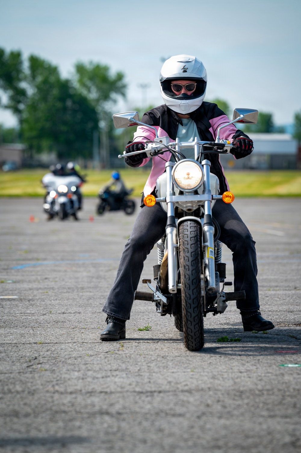 A rider wearing a white full-face helmet and pink-and-black jacket is operating a silver motorcycle on a paved training course. Both feet are planted on the ground, suggesting the motorcycle is stopped or moving very slowly. In the background, two additional riders on separate motorcycles are visible, along with orange traffic cones marking the course layout.