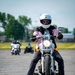 A rider wearing a white full-face helmet and pink-and-black jacket is operating a silver motorcycle on a paved training course. Both feet are planted on the ground, suggesting the motorcycle is stopped or moving very slowly. In the background, two additional riders on separate motorcycles are visible, along with orange traffic cones marking the course layout.