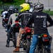 A group of motorcyclists are lined up in a parking lot, likely participating in a motorcycle safety or training course. Riders are wearing helmets and seated on various motorcycles, facing forward in a queue. Orange traffic cones are visible in the background, suggesting a structured riding exercise.