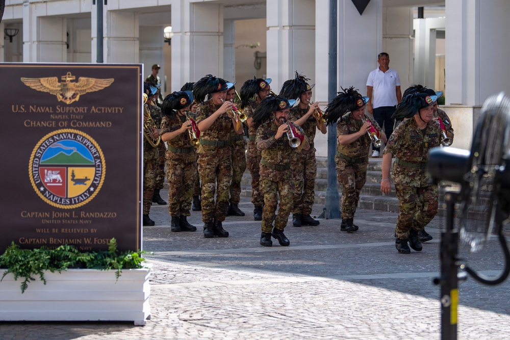 A military band in camouflage uniforms and distinctive black feathered hats is performing at a U.S. Naval Support Activity Change of Command ceremony in Naples, Italy. The sign in the foreground indicates that Captain John L. Randazzo, United States Navy, is being relieved by Captain Benjamin W. Foster. The event is taking place outdoors on a cobblestone plaza in front of a white colonnaded building.