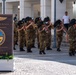 A military band in camouflage uniforms and distinctive black feathered hats is performing at a U.S. Naval Support Activity Change of Command ceremony in Naples, Italy. The sign in the foreground indicates that Captain John L. Randazzo, United States Navy, is being relieved by Captain Benjamin W. Foster. The event is taking place outdoors on a cobblestone plaza in front of a white colonnaded building.