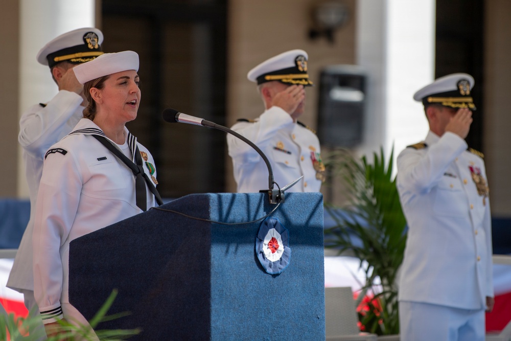 A U.S. Navy enlisted sailor in dress whites is speaking at a podium during a formal military ceremony. Behind her, two senior naval officers in dress white uniforms with gold insignia are rendering a salute. The setting appears to be an outdoor ceremonial venue with classical architectural columns in the background.