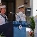 A U.S. Navy enlisted sailor in dress whites is speaking at a podium during a formal military ceremony. Behind her, two senior naval officers in dress white uniforms with gold insignia are rendering a salute. The setting appears to be an outdoor ceremonial venue with classical architectural columns in the background.