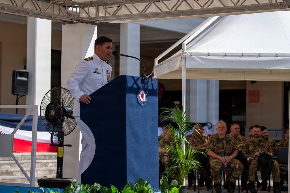 A naval officer in white dress uniform is delivering a speech at a formal military ceremony from a blue podium with an emblem. Several military personnel in camouflage uniforms are seated in the background as an audience. The event appears to be an outdoor ceremony held under a temporary canopy structure.