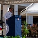 A naval officer in white dress uniform is delivering a speech at a formal military ceremony from a blue podium with an emblem. Several military personnel in camouflage uniforms are seated in the background as an audience. The event appears to be an outdoor ceremony held under a temporary canopy structure.
