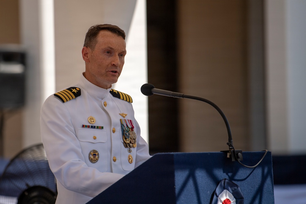 A naval officer in white dress uniform is speaking at a podium equipped with a microphone. The officer displays multiple medals, ribbons, and insignia on their uniform, indicating significant military service and rank. The setting appears to be a formal military ceremony or official event.