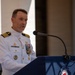 A naval officer in white dress uniform is speaking at a podium equipped with a microphone. The officer displays multiple medals, ribbons, and insignia on their uniform, indicating significant military service and rank. The setting appears to be a formal military ceremony or official event.
