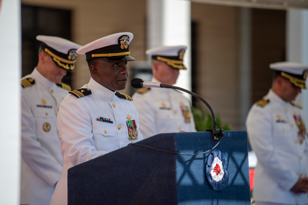 A naval officer in white dress uniform is speaking at a podium during what appears to be a formal military ceremony. Several other naval officers in white dress uniforms stand in the background. The podium features a blue covering with an emblem that includes a red cross symbol.