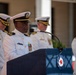 A naval officer in white dress uniform is speaking at a podium during what appears to be a formal military ceremony. Several other naval officers in white dress uniforms stand in the background. The podium features a blue covering with an emblem that includes a red cross symbol.
