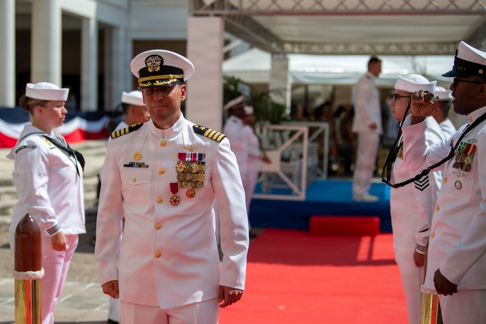 A formal U.S. Navy military ceremony is taking place outdoors, featuring personnel in white dress uniforms. A senior officer, likely a Captain or Admiral based on rank insignia and numerous medals, stands prominently in the foreground on a red carpet. Supporting naval personnel are positioned in formation, with one sailor playing a ceremonial instrument on the right.