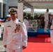 A formal U.S. Navy military ceremony is taking place outdoors, featuring personnel in white dress uniforms. A senior officer, likely a Captain or Admiral based on rank insignia and numerous medals, stands prominently in the foreground on a red carpet. Supporting naval personnel are positioned in formation, with one sailor playing a ceremonial instrument on the right.