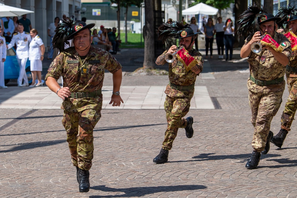 Italian Army (Esercito) soldiers in camouflage uniforms and distinctive black feathered hats are marching in a military parade or ceremony. Two of the soldiers are playing trumpets adorned with red and gold decorative banners featuring the Italian flag. The event appears to take place in an urban plaza with civilian spectators and naval officers visible in the background.