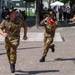 Italian Army (Esercito) soldiers in camouflage uniforms and distinctive black feathered hats are marching in a military parade or ceremony. Two of the soldiers are playing trumpets adorned with red and gold decorative banners featuring the Italian flag. The event appears to take place in an urban plaza with civilian spectators and naval officers visible in the background.