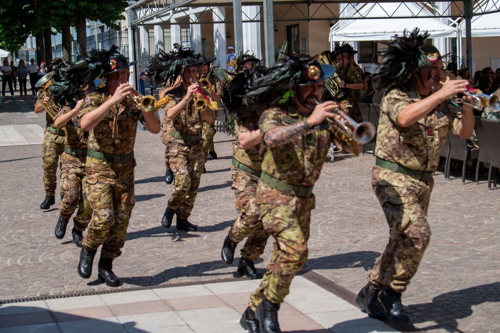 A military marching band is performing in an outdoor public square, with musicians wearing camouflage uniforms and distinctive black feathered hats. The performers are playing brass instruments, primarily trumpets, while marching in formation. The setting appears to be a European town square with spectators and outdoor café seating visible in the background.