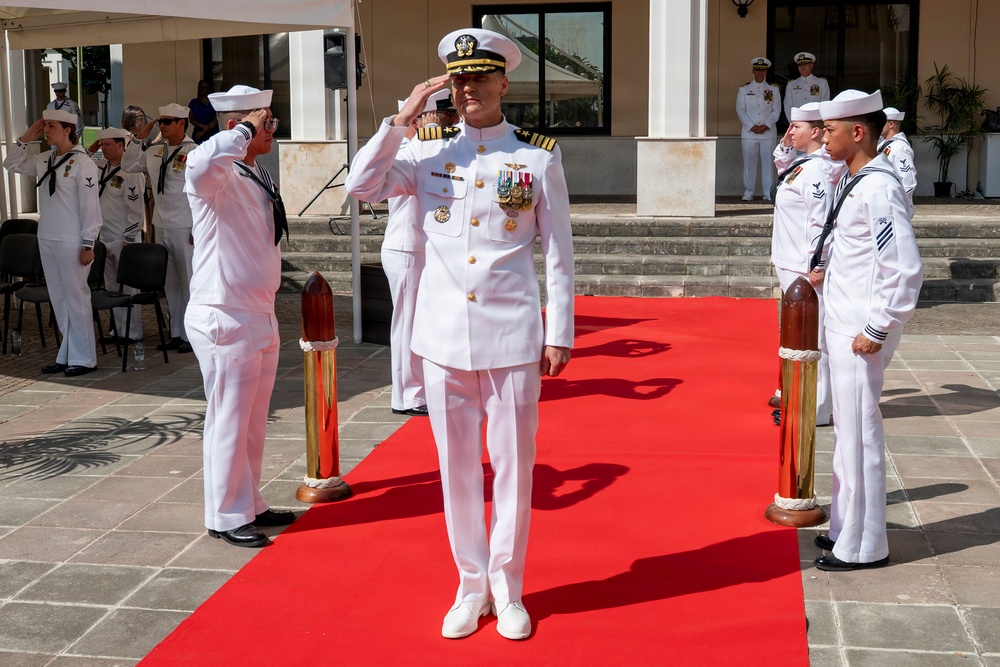 A formal U.S. Navy military ceremony is taking place outdoors on a red carpet, likely a change of command or official reception. A senior officer in white dress uniform with captain's insignia and multiple medals stands at the center saluting, flanked by sideboys standing at attention along the red carpet. Additional naval personnel in white uniforms are visible in the background observing the ceremony.