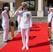 A formal U.S. Navy military ceremony is taking place outdoors on a red carpet, likely a change of command or official reception. A senior officer in white dress uniform with captain's insignia and multiple medals stands at the center saluting, flanked by sideboys standing at attention along the red carpet. Additional naval personnel in white uniforms are visible in the background observing the ceremony.