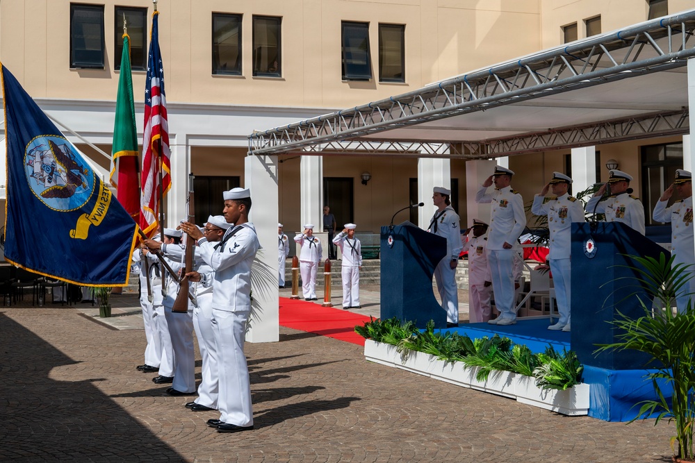 A formal U.S. Navy military ceremony is taking place in an outdoor courtyard, featuring a color guard presenting flags including the American flag, a green flag (likely Portuguese), and a Navy unit flag. Officers in white dress uniforms are saluting on a raised platform with a podium, while enlisted personnel stand at attention. The setting appears to be a naval installation, possibly in a Mediterranean or Portuguese-speaking country based on the architectural style and flag presence.