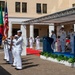 A formal U.S. Navy military ceremony is taking place in an outdoor courtyard, featuring a color guard presenting flags including the American flag, a green flag (likely Portuguese), and a Navy unit flag. Officers in white dress uniforms are saluting on a raised platform with a podium, while enlisted personnel stand at attention. The setting appears to be a naval installation, possibly in a Mediterranean or Portuguese-speaking country based on the architectural style and flag presence.
