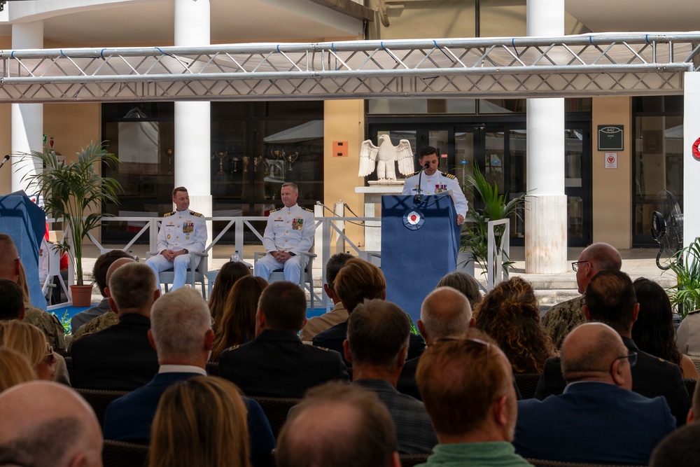A formal military ceremony is taking place outdoors in a courtyard setting, with one officer speaking at a podium while two other uniformed officers are seated on a stage behind him. The audience consists of numerous civilians and some military personnel seated in rows facing the stage. The setting appears to be a military installation, likely a change of command or similar official ceremony.