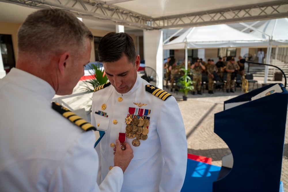 A military decoration ceremony is taking place outdoors, where a senior naval officer is pinning a medal onto the uniform of another officer in white dress uniform. The recipient is wearing an extensive collection of medals and decorations on his chest. The ceremony appears to be formal, with additional military personnel visible in the background.