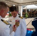 A military decoration ceremony is taking place outdoors, where a senior naval officer is pinning a medal onto the uniform of another officer in white dress uniform. The recipient is wearing an extensive collection of medals and decorations on his chest. The ceremony appears to be formal, with additional military personnel visible in the background.