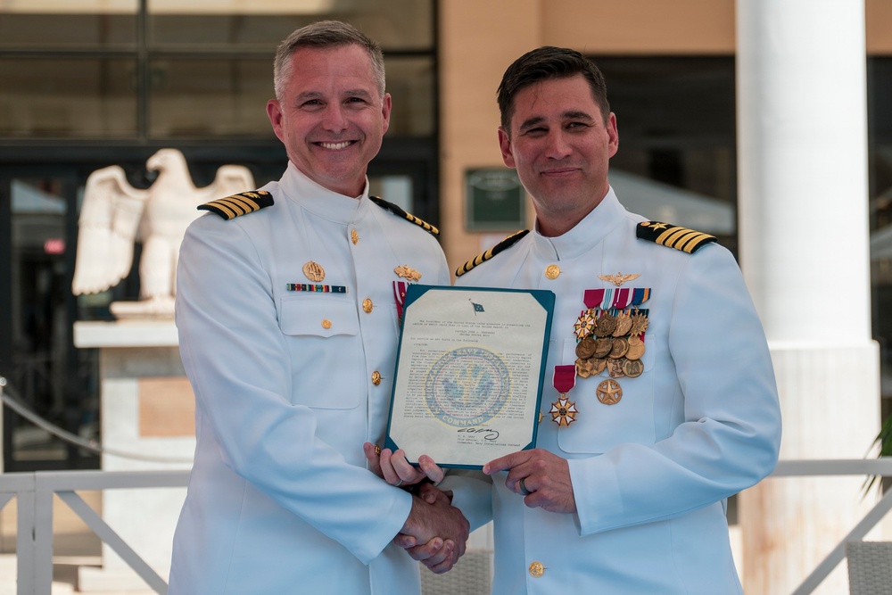 Two U.S. Navy officers in white dress uniforms are posing together while holding an official document, likely a commission or award certificate. The officer on the right displays an extensive array of medals and decorations on his chest, suggesting a promotion or retirement ceremony. Both officers are shaking hands in a formal presentation pose outdoors.