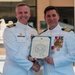 Two U.S. Navy officers in white dress uniforms are posing together while holding an official document, likely a commission or award certificate. The officer on the right displays an extensive array of medals and decorations on his chest, suggesting a promotion or retirement ceremony. Both officers are shaking hands in a formal presentation pose outdoors.