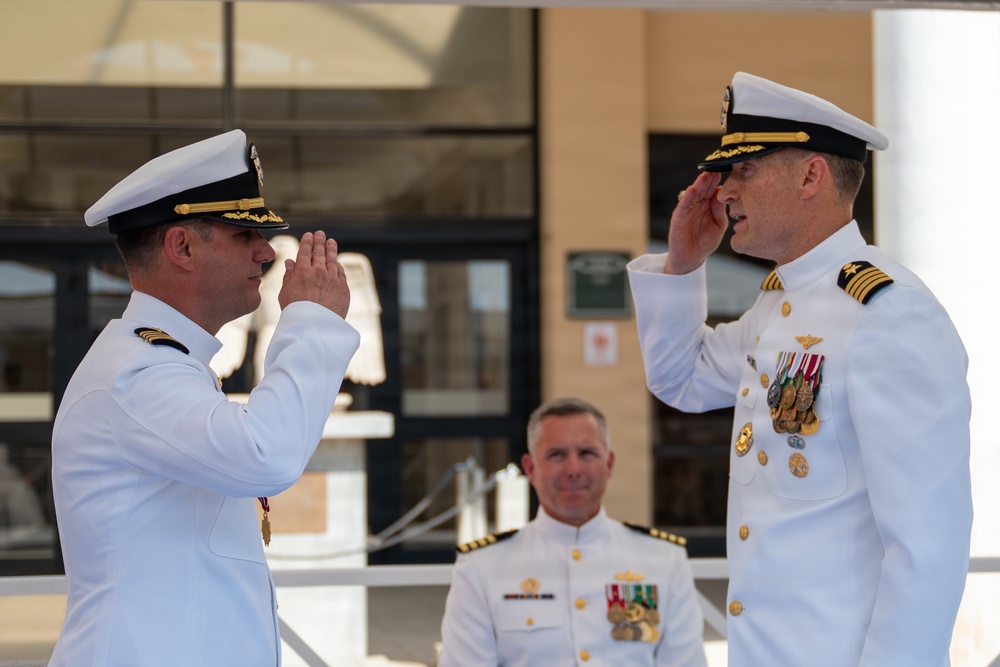 Two U.S. Navy officers in white dress uniforms are exchanging salutes in what appears to be a formal military ceremony, likely a change of command or retirement event. A third Navy officer stands in the background observing the exchange. The setting appears to be outdoors in front of a military building.