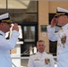Two U.S. Navy officers in white dress uniforms are exchanging salutes in what appears to be a formal military ceremony, likely a change of command or retirement event. A third Navy officer stands in the background observing the exchange. The setting appears to be outdoors in front of a military building.