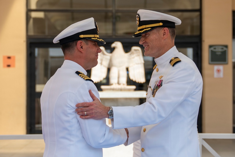 Two U.S. Navy officers in white dress uniforms are engaged in a formal greeting or congratulatory exchange outside a military building. The officer on the right displays multiple service ribbons and medals, suggesting higher seniority or longer service. A decorative eagle sculpture is visible in the background through glass doors.