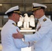 Two U.S. Navy officers in white dress uniforms are engaged in a formal greeting or congratulatory exchange outside a military building. The officer on the right displays multiple service ribbons and medals, suggesting higher seniority or longer service. A decorative eagle sculpture is visible in the background through glass doors.